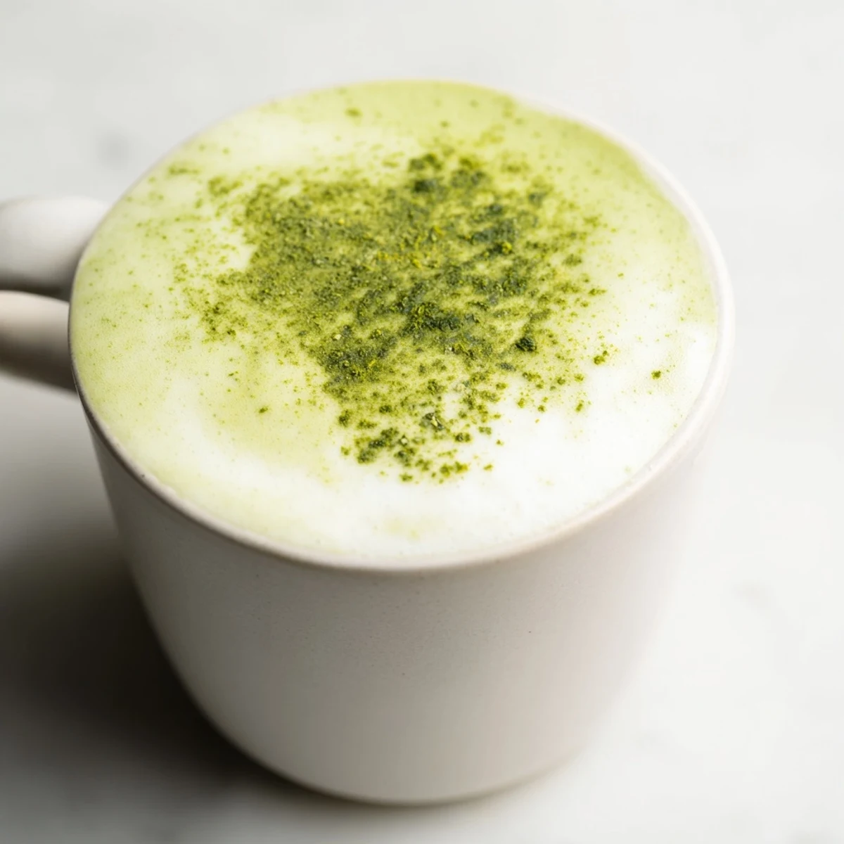 Creamy green matcha latte with almond milk in a white ceramic mug, topped with light froth and a dusting of matcha powder.