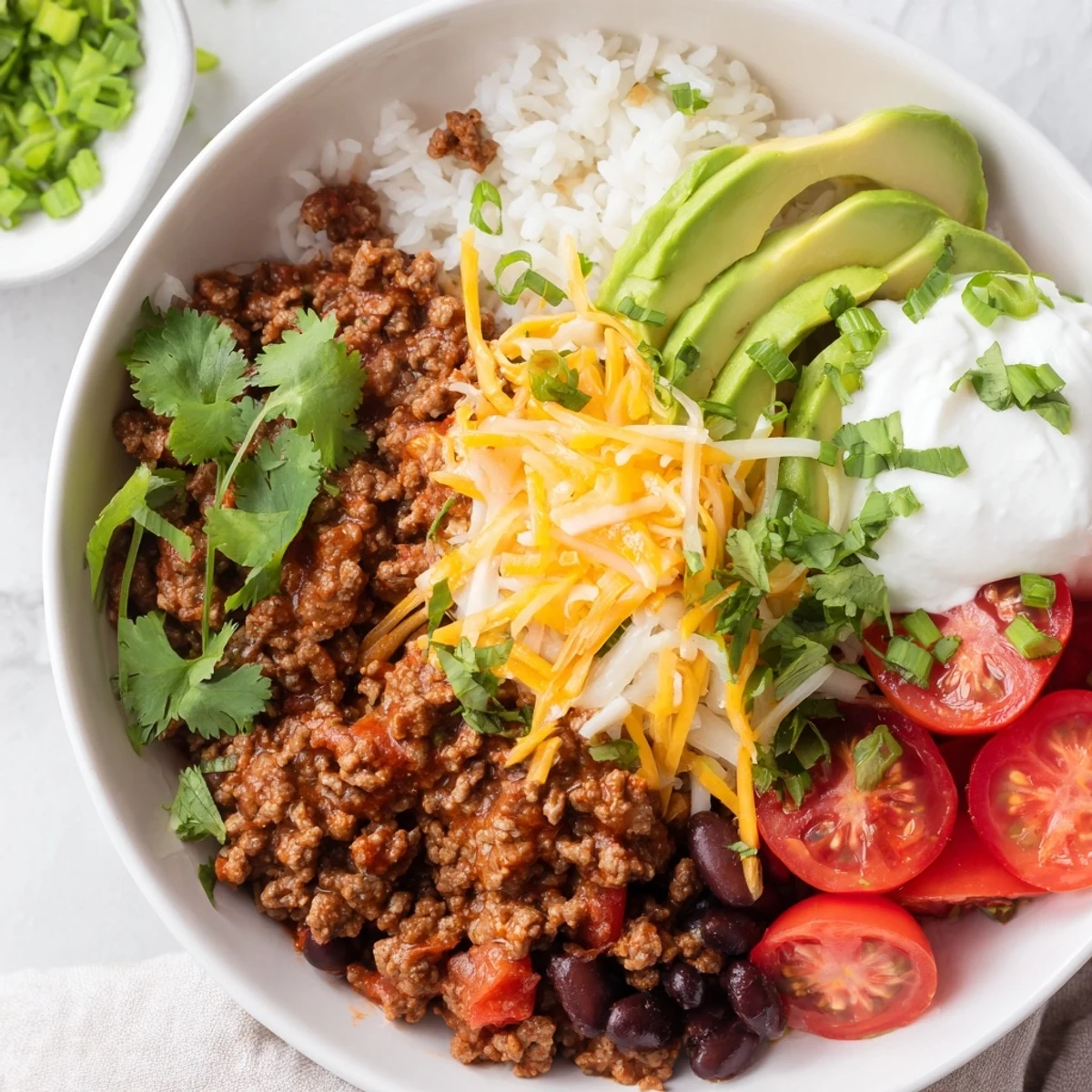 A close-up of Beef Burrito Bowls with Rice and Beans topped with seasoned ground beef, black beans, avocado slices, shredded cheese, sour cream, and fresh cilantro. 